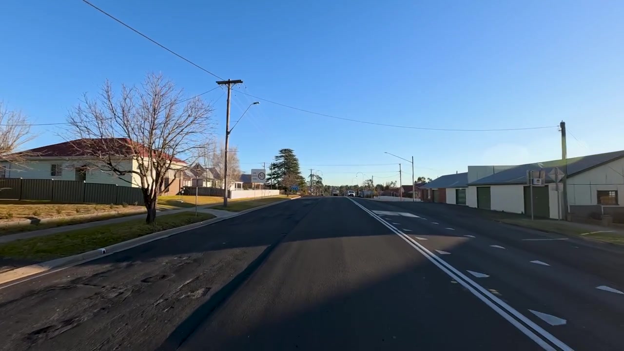 Et kjøretøy kjører langs en solrik asfaltvei forbi mursteinsbygninger i Uralla, Australia