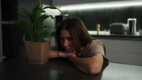 una chica morena triste y pensativa en una camiseta beige se sienta apoyada en una mesa de comedor negra en la cocina cerca de una planta verde de interior en una olla por la noche en una cocina moderna