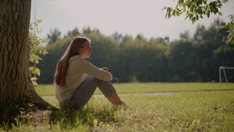 thoughtful young woman sits on grassy ground with folded arms over her knees, gazing into distance as sunlight reflects softly on her face, with a blurred background of stadium and poles