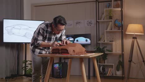 asian man automotive designer having a headache while using spatula to smooth out the surface and create details in the sculpture of car clay in the studio