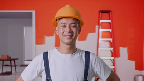 close up de hombre asiático pintor con casco de seguridad de pie con los brazos akimbo mirando a la cámara y sonriendo con la pared de pintura en la habitación