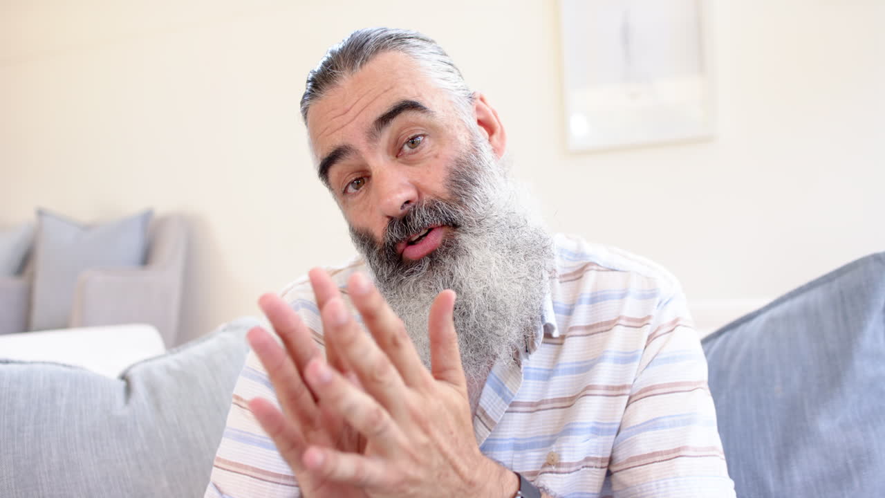 Senior man with gray beard talking and gesturing while sitting on couch