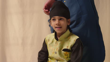 studio-aufnahme eines vaters, der einen turban auf den kopf eines jungen sikh-sohnes bindet, der eine traditionelle weste trägt.