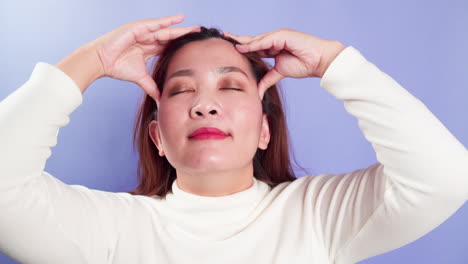 보라색 배경 위에 고립된 자기 머리 마사지로 두통 편두통 개념에 대처하는 좌절된 스트레스를 받은 젊은 아시아 여성
