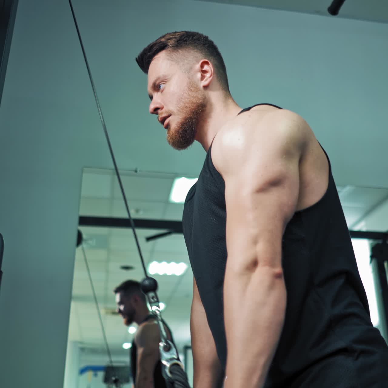 Side view of fit sportsman training his muscles. Muscular athlete is pulling rope on the modern simulator in sports center. Close-up.