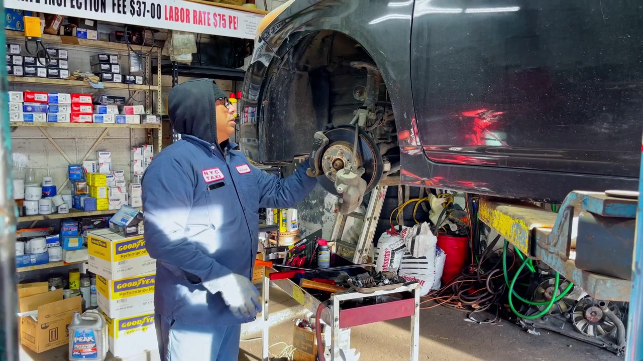 2022年3月9日、アメリカ合衆国ニューヨーク：車のフロントブレーキキャリパーを点検するプロの自動車整備士。作業場でリフトアップされた車のブレーキディスクアセンブリを点検する、NYCタクシージャケットを着た自動車技術者。
