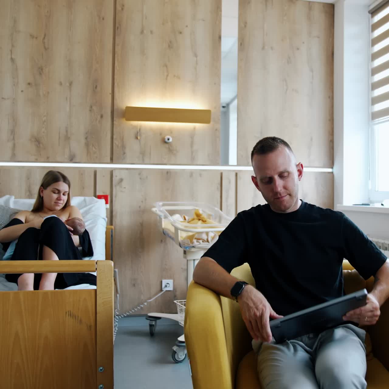 New-made Caucasian parents are in the hospital ward after childbirth. Man sits to the armchair with computer and woman breastfeeding her child