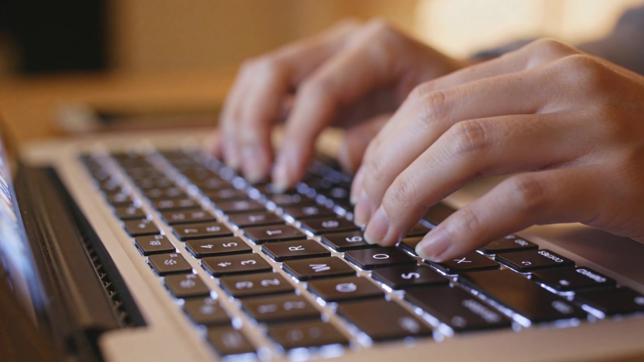 Typing hands in gray sleeves with fingers poised starting keystrokes at desk, backlit keyboard