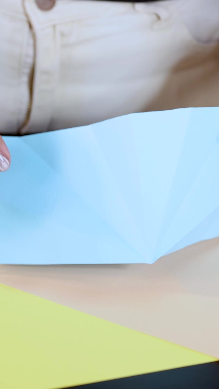 Person folds blue paper on table, demonstrating origami technique in bright, natural lighting