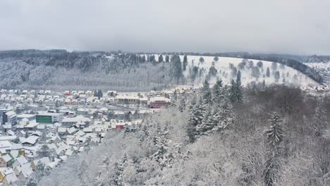 Pullback-winter-shot,-flying-backwards-to-discover-a-snowy-city-behind-a-hill-with-snowy-white-trees-at-the-recreation-area-of-the-Swabian-Alb