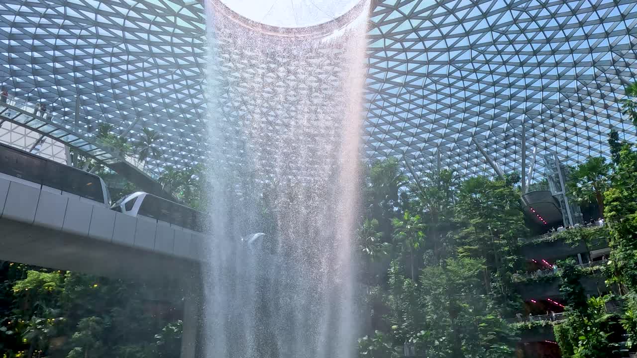 Monorail passes lush greenery as indoor waterfall cascades under glass dome, natural daylight, wide shot
