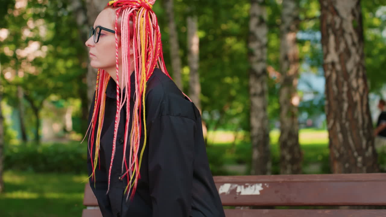 Mujer caucásica con trenzas de neón, inclinada hacia delante en un banco de madera en un bosque de abedules, con las manos cerca de la cara y expresión de preocupación, parque iluminado por el sol de fondo y ambiente introspectivo, esperando noticias