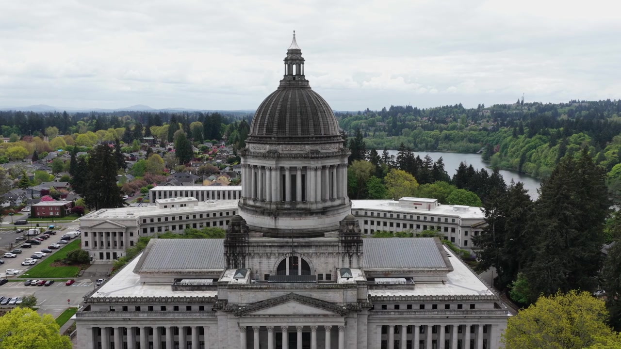 Campidoglio dello Stato di Washington a Olympia con storica cupola in pietra ed edifici legislativi che si affacciano sul Lago Capitol con alberi verdi lussureggianti e auto nel parcheggio sotto un cielo nuvoloso. Ampia ripresa aerea laterale.