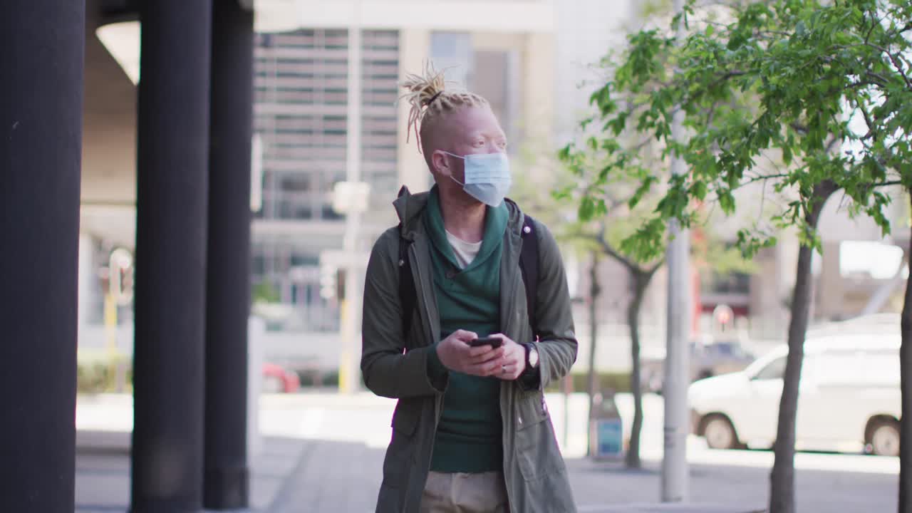 Albino african american man with face mask and dreadlocks walking and using smartphone