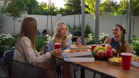 a blonde girl eats pizza with her friends and chats while relaxing at a table in the courtyard of a country house