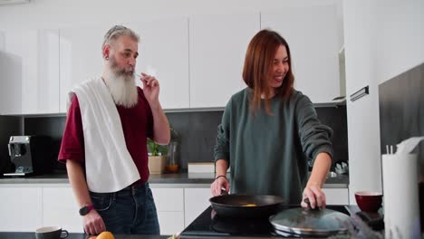 Happy-brunette-girl-in-a-green-sweater-prepares-breakfast-while-her-father-an-elderly-man-with-gray-hair-and-a-lush-beard-brushes-his-teeth-in-the-kitchen-in-a-modern-apartment