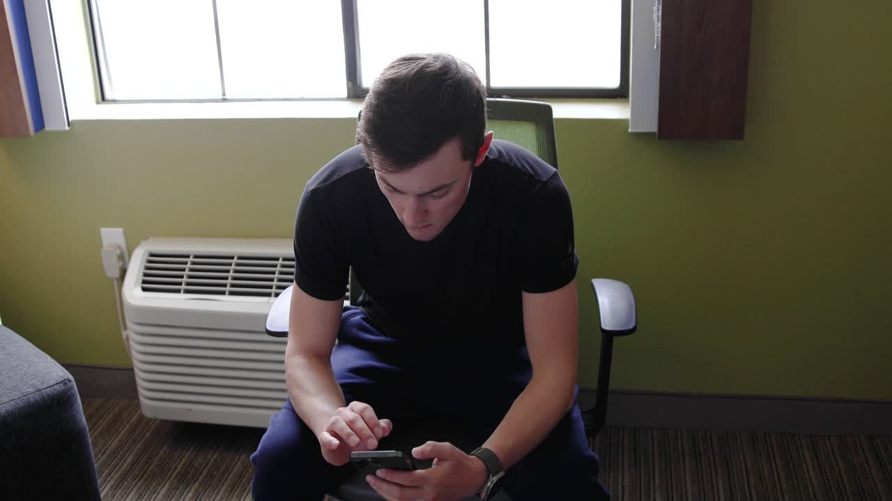 Male on a chair in front of a white heater texting on his phone in a hotel room