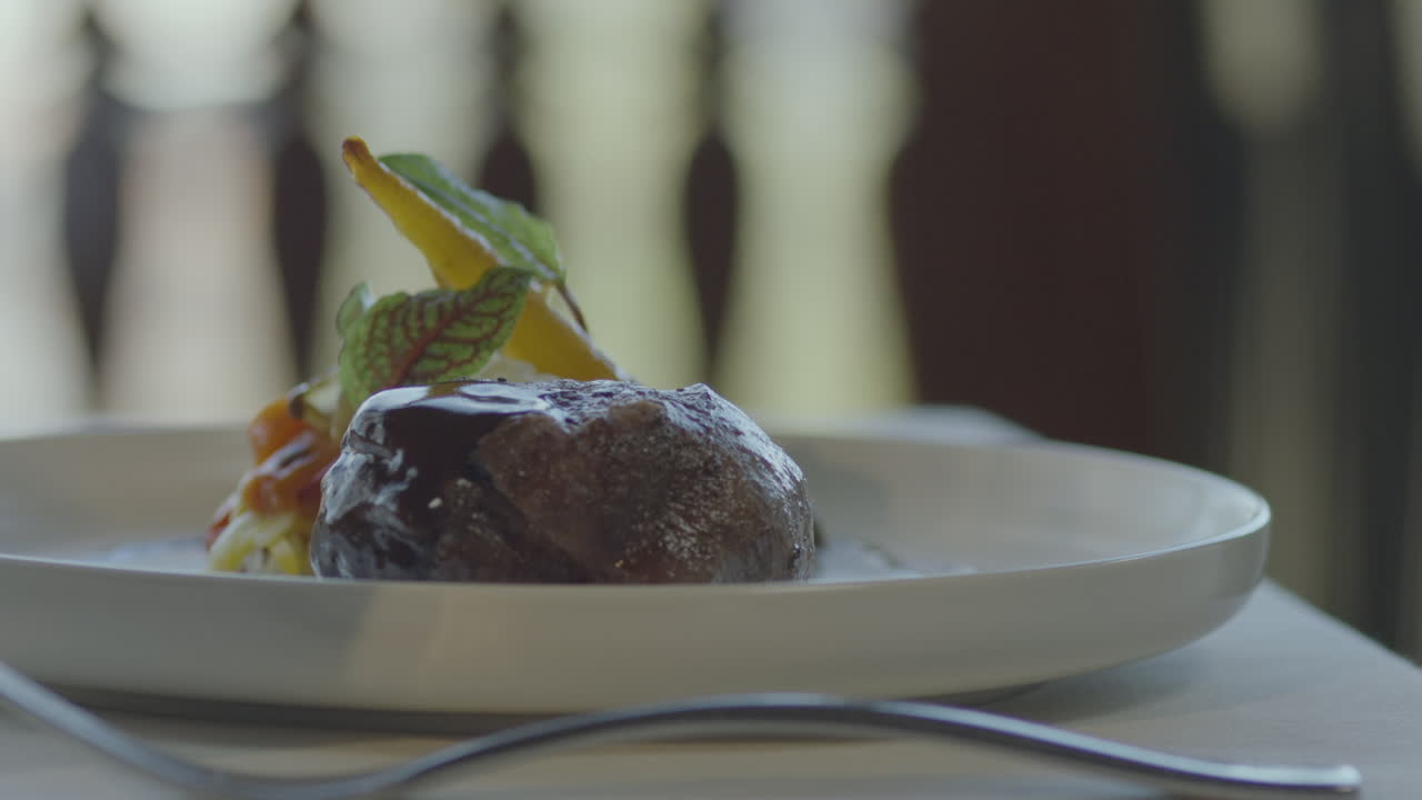 Close-up of a gourmet dish served on a white plate: meat covered with a light glaze, green leaves and mini vegetables on one side