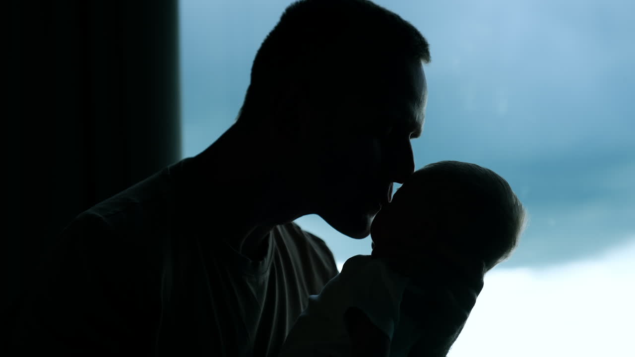 Dark male silhouette holding a baby close to face. Caring dad kisses his child on the cheek.