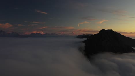 Drone-shot-flying-over-the-edge-of-a-cliff-on-top-of-a-mountain,-revealing-another-peak-surrounded-by-soft-and-white-clouds-at-extreme-altitudes-on-Mount-Niesen,-Switzerland-during-a-sunrise-or-sunset