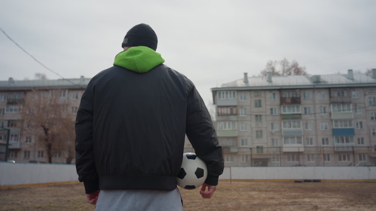 male strolls confidently, man with football outdoors, young man carrying soccer ball in muted weather, person dressed in hoodie demonstrating calm assurance while moving across field