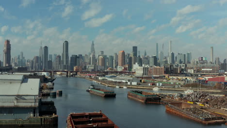 aerial-descending-footage-of-ship-pushing-barge-on-Newtown-Creek.-Cargo-boats-moored-at-banks.-Skyline-with-skyscrapers.-New-York-City,-USA