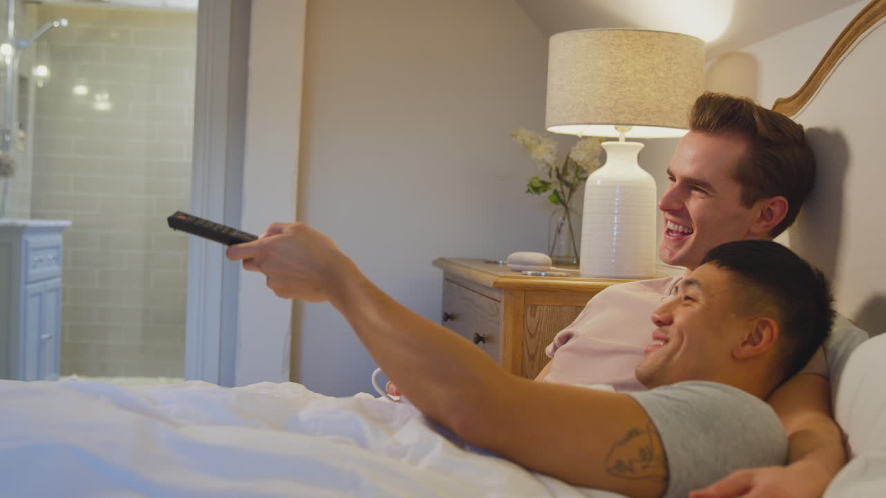 Coppia di uomini innamorati sdraiata a letto a casa che guarda la TV insieme