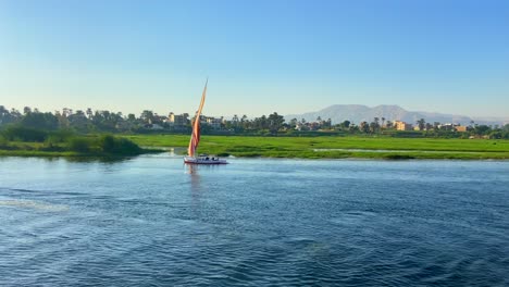 Filmische-4K-Aufnahmen-In-Zeitlupe-Eines-ägyptischen-Segelboots-Und-Eines-Kleinen-Motorschiffs,-Das-Bei-Sonnenuntergang-Am-Ufer-Des-Niltals-Entlang-Segelt