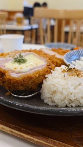 crispy cheese-filled tonkatsu sliced open, served with steamed rice on a plate in cozy korean restaurant setting, revealing woman eating korean woman blurred in backdrop - parallax close-up vertical