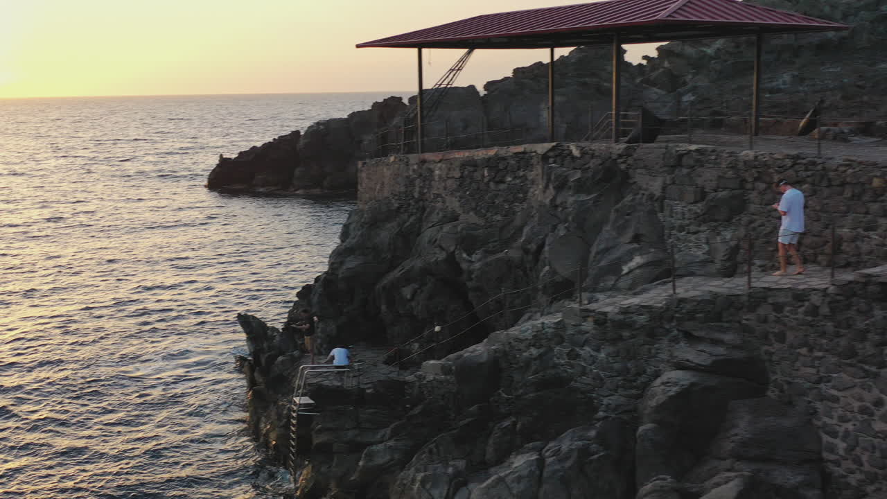 Guy walking down from a stone pier to a cliff above the sea,flying a drone