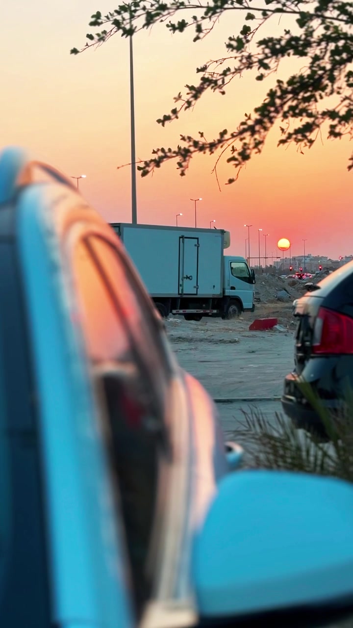 A cinematic vertical shot of a white box truck parked in an outdoor lot during a golden sunset. A blurred blue car rests in the foreground beneath tree branches in the warm evening.