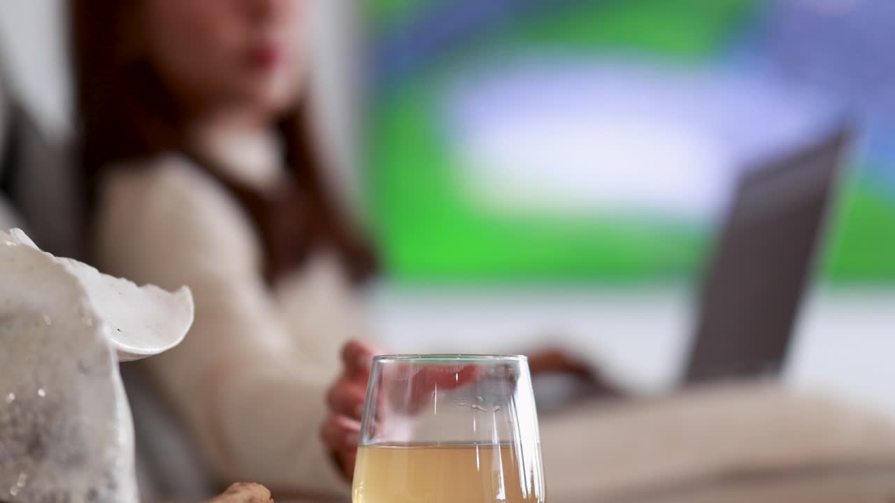 An Asian woman sits on a sofa with a laptop, drinking juice and watching television in a softly lit, modern living room. Shallow depth of field emphasizes relaxation