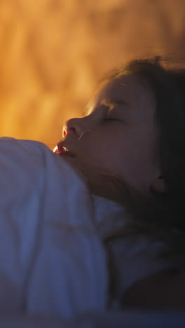 little girl silhouette covered with warm blanket sleeps on comfortable bed in semi-dark room with glowing night lamp resting at home closeup slow motion