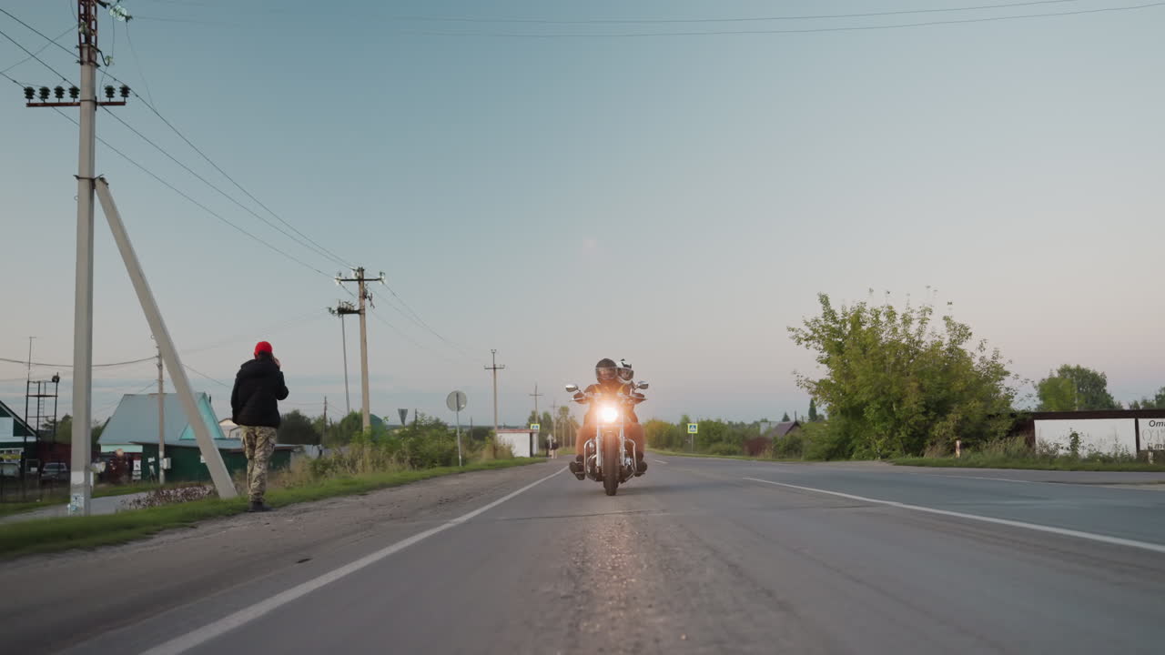 Motorcycle rider with passenger wearing helmets rides countryside road during trip, passing pedestrians on roadside with utility poles, houses, trees, and clear evening sky