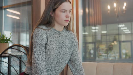 College student bending over to place cold drink on table, sits comfortably in mall cafe, lifts head and looks around relaxed. Background shows chairs, tables, glass mirrors, indoor plants
