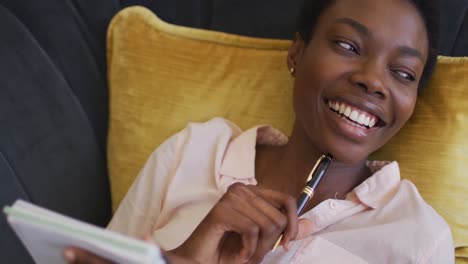 Feliz-Mujer-Afroamericana-Tumbada-En-El-Sofá-De-La-Sala-De-Estar,-Escribiendo-En-Un-Cuaderno