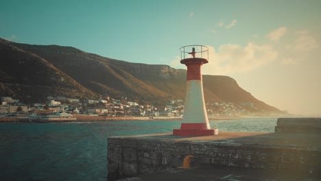 Small-lighthouse-on-the-coast,-beautiful-coastal-village-located-on-the-hills-in-background