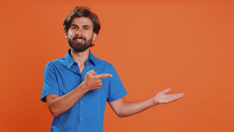 hombre en camisa azul señalando y sonriendo, de pie frente a un fondo naranja