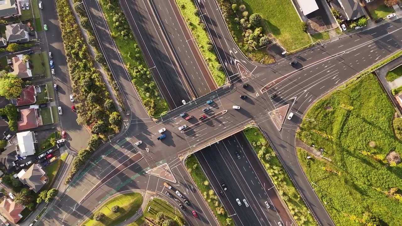 Top-down aerial view of highways crossing through Mount Roskill, Auckland, with marked lanes, turning routes, vehicles, landscaped areas, and surrounding homes in warm evening light.