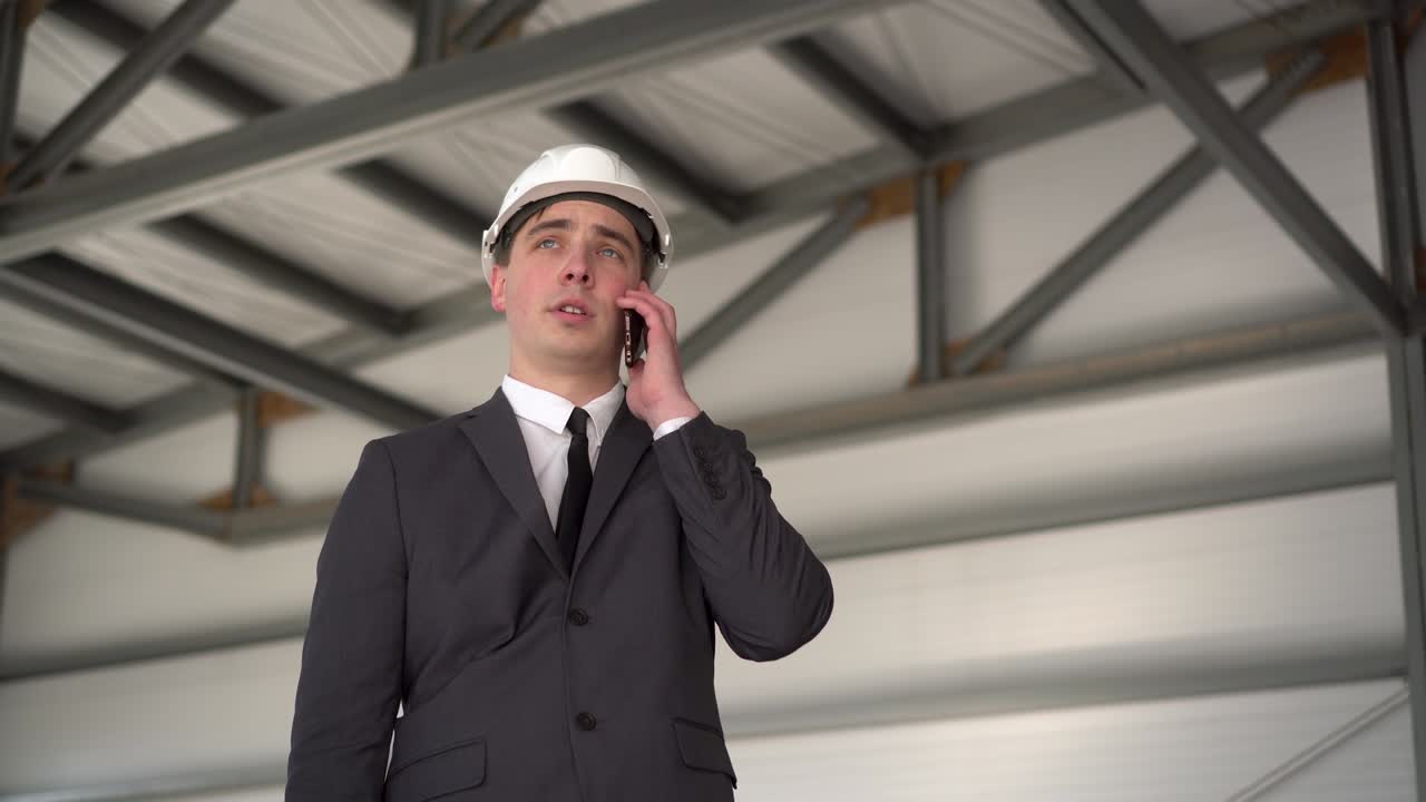 A young man in a helmet speaks on the phone at a construction site. The boss in a suit is talking on the phone.