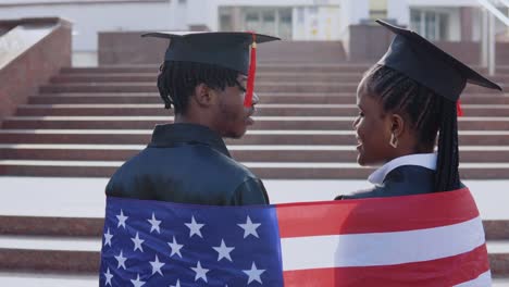 african american man and woman standing side by side with their backs to the camera. they have the british flag on their shoulders. a university building on background