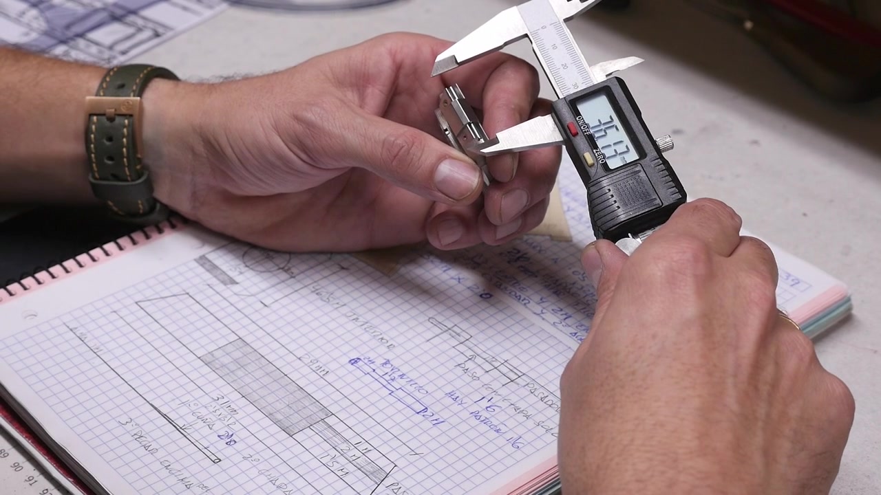 Primo piano di mani artigiane che usano un calibro a corsoio elettronico per misurare una parte di orologio sopra un quaderno con disegni tecnici, schizzi e modelli in pelle.
