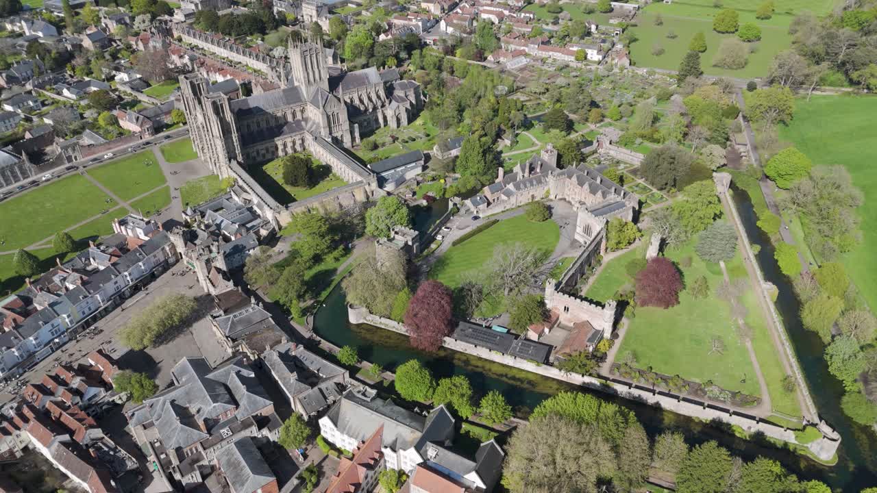 Orbital aerial view of medieval cathedral complex with soaring gothic spires, adjacent bishop’s palace, and surrounding historic townscape amid lush gardens and lawns