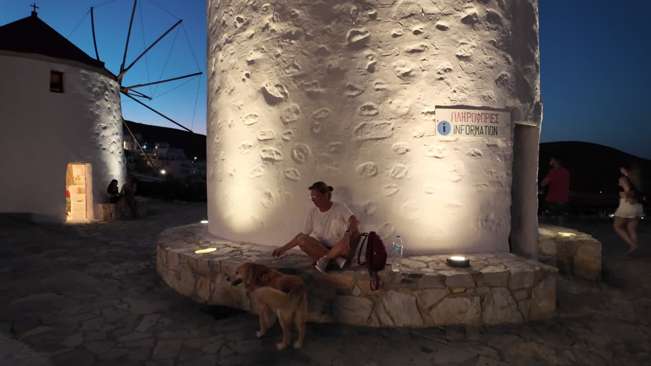Greece, Astypalea Island, iconic windmills nicely lighted up at night time. A woman sitting by the wind mill and her golden retriever is around her moving slowly. Camera moves slowly to the blue sky.