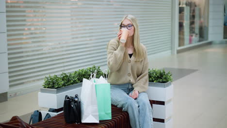 lady with long hair drinking coffee from a cup while wearing knitted sweater, sitting in a mall, with clothing store in background showcasing stylish garments and accessories