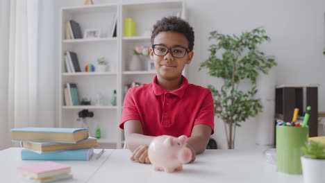 african american kid putting coin in piggybank, looking at camera, deposit