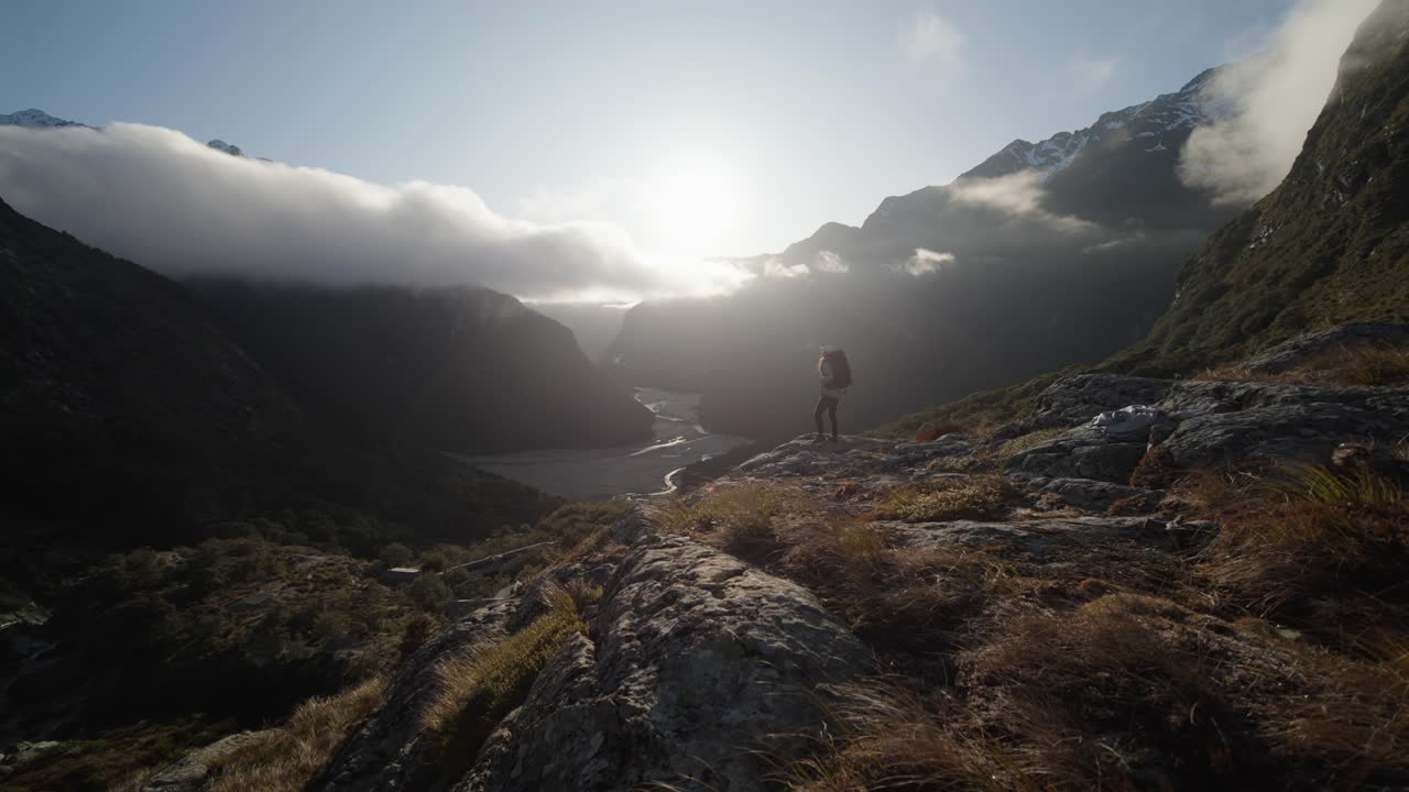 A girl hiking and exploring mountains and valleys on a beautiful morning during sunrise. I'ts a calm quiet day. Routeburn Hike, New Zealand. Handheld very wide shot.