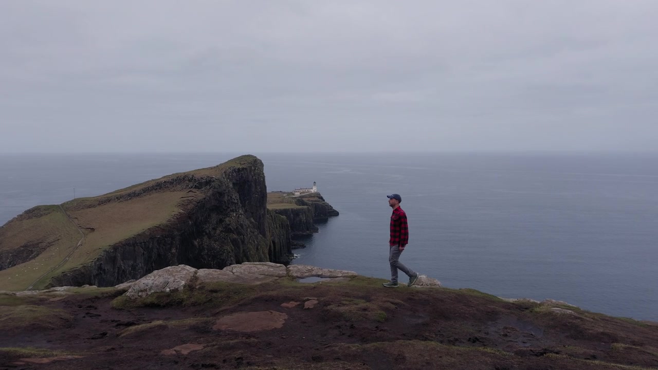 Persona in piedi sul bordo di una scogliera che si affaccia sull'oceano sotto un cielo nuvoloso nella Scozia remota, con il faro di Neist Point sullo sfondo.