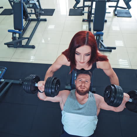 portrait of athletic man with muscular body lifting dumbbells in gym. strong man lifts heavy dumbbells with the help of beautiful red-haired woman in sport club indoors. top view.