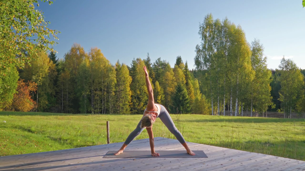 Fit caucasian woman in sportswear practicing yoga stretches, the wide-legged forward fold with twist on a yoga mat outdoors in forest on a wooden platform, rotating her spine and stretching hamstrings